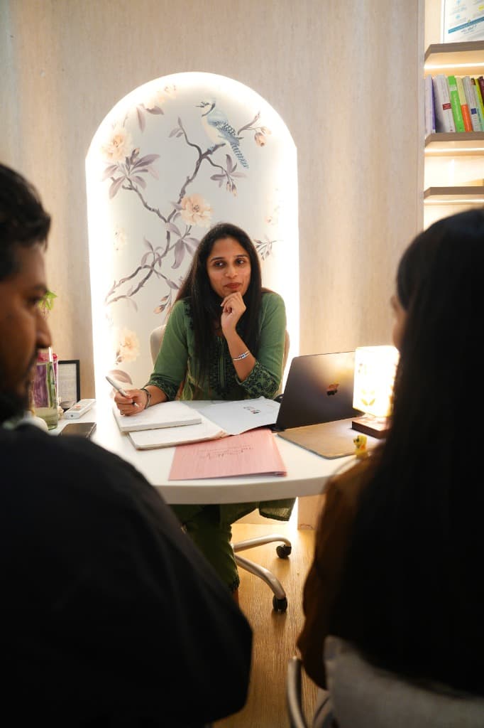 A woman and a man sit side by side at a wooden table, talking with a female clinician who holds a pen above an open notebook.